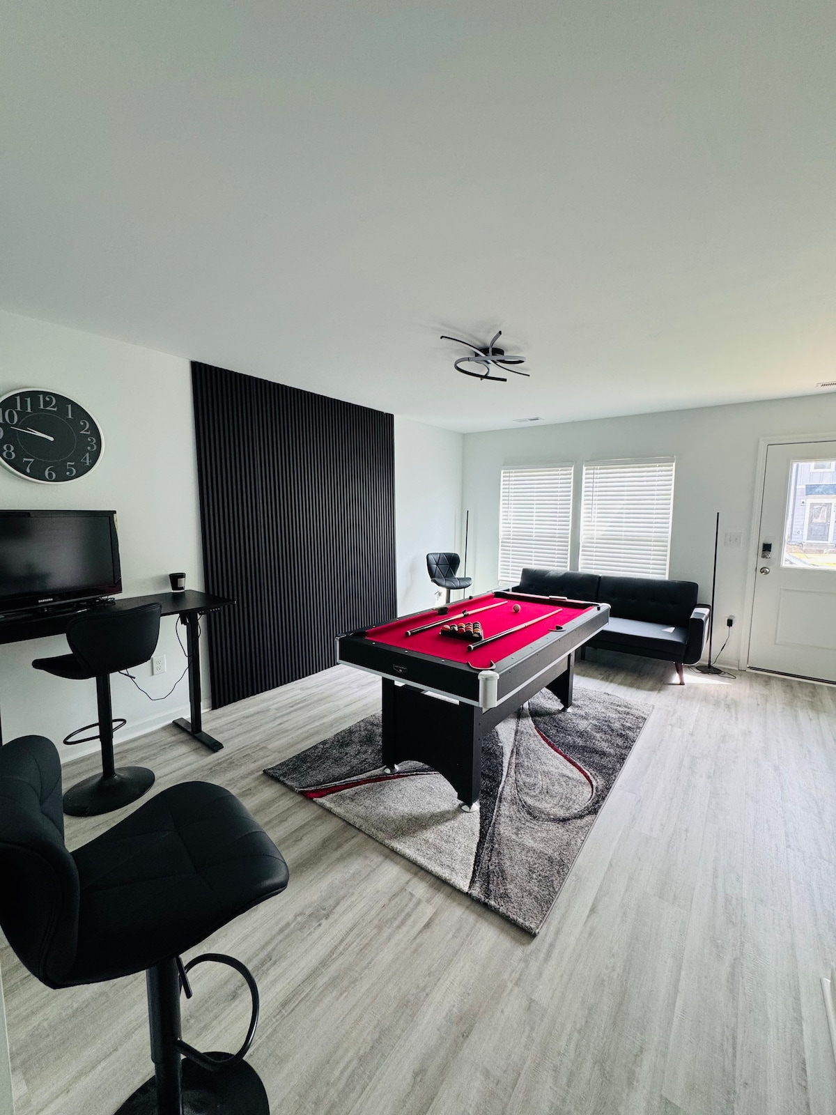 A vibrant game room is featured, showcasing a pool table with a red felt surface at its center. Two seating areas, including a black sofa and bar stools, are arranged around a modern rug. Natural light streams in through the nearby windows.