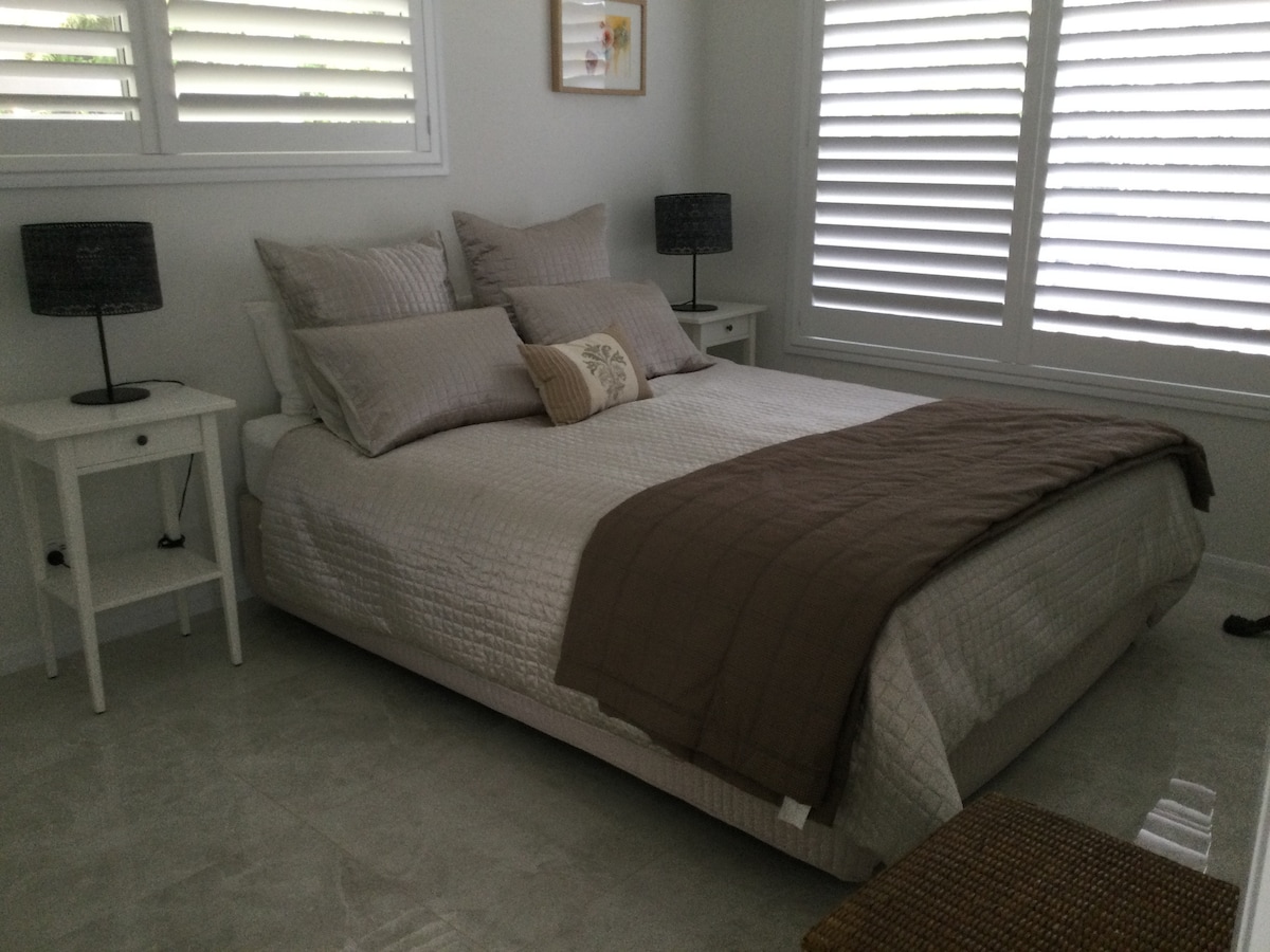 A spacious bed is dressed in soft, neutral linens, accented by decorative pillows. Each side of the bed features a nightstand with a lamp. Natural light filters through the white plantation shutters, illuminating the serene atmosphere of the room.