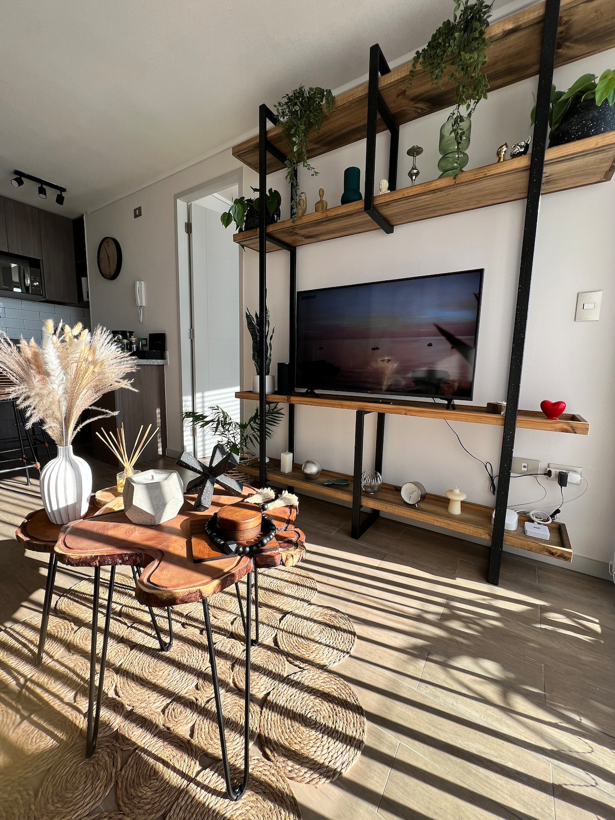 A modern living area features an arrangement of wooden shelves adorned with plants and decorative items. A flat-screen TV is mounted on the wall, while a low table made of sliced wood with black legs is positioned in front, complemented by a circular woven rug beneath.