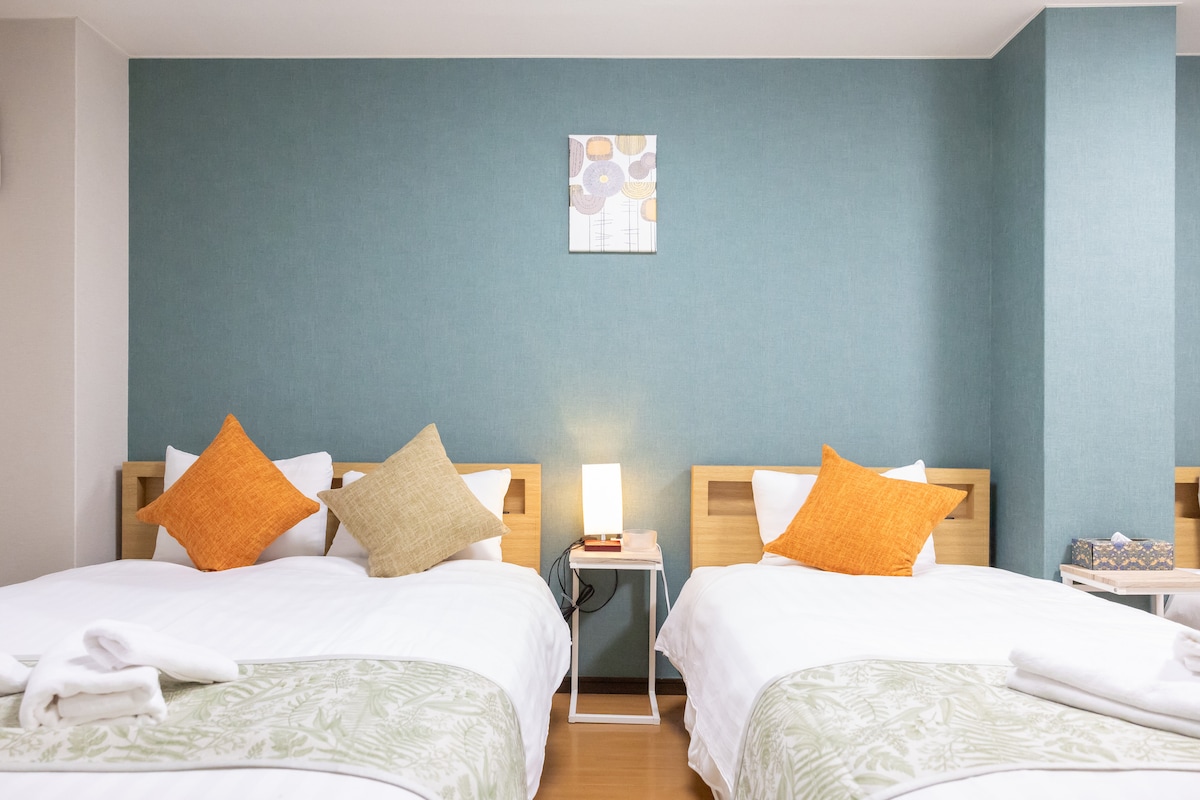 The room features two spacious beds with light linens, accented by decorative pillows in warm hues. A bedside table holds a lamp and a small book. A piece of wall art is displayed above, set against a calming blue wall.