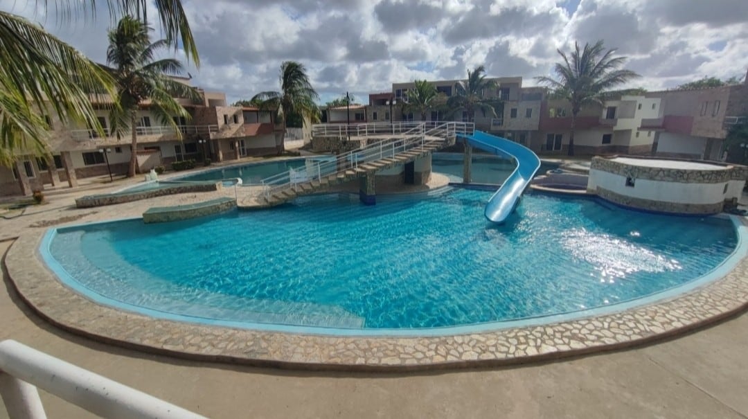 A spacious pool area features a large swimming pool with a winding water slide, surrounded by palm trees. A bridge crosses over the pool, providing access to different sections. The clear blue water is complemented by a paved walkway along the edge.