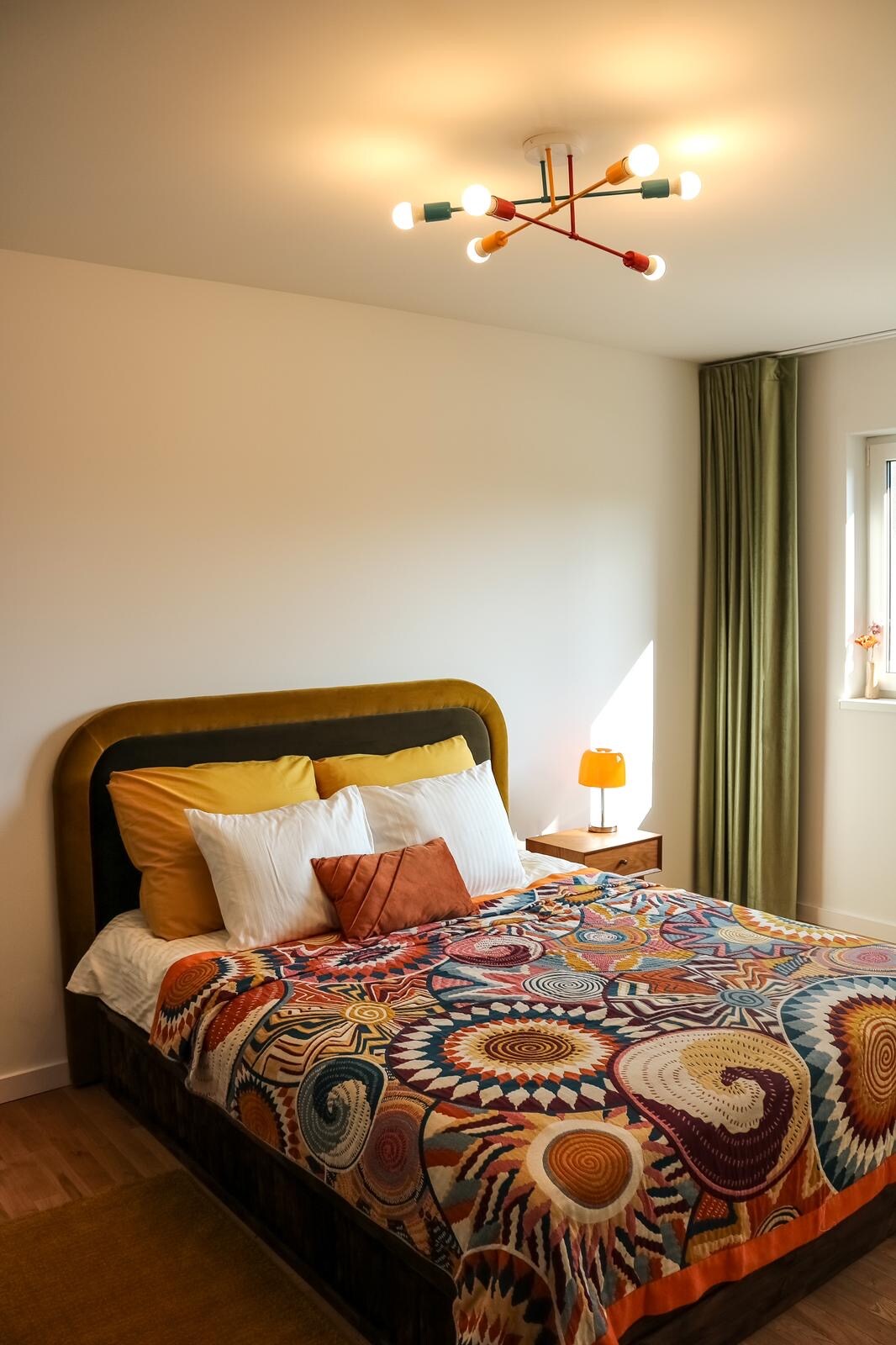 A comfortable bedroom features a spacious bed adorned with a colorful patterned blanket and numerous pillows. A bedside table with an orange lamp complements the overall design. Light enters through a window dressed with green curtains, enhancing the room's bright and welcoming atmosphere.