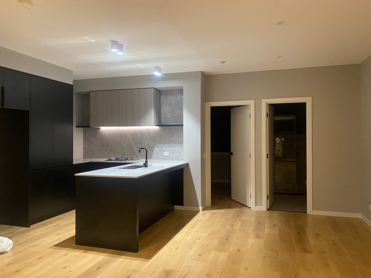 A modern kitchen area is highlighted by a central island with a light-colored countertop. Dark cabinetry contrasts with lighter wall panels. Soft lighting is provided by recessed fixtures, creating a bright but neutral atmosphere. An open doorway leads to an adjacent room.
