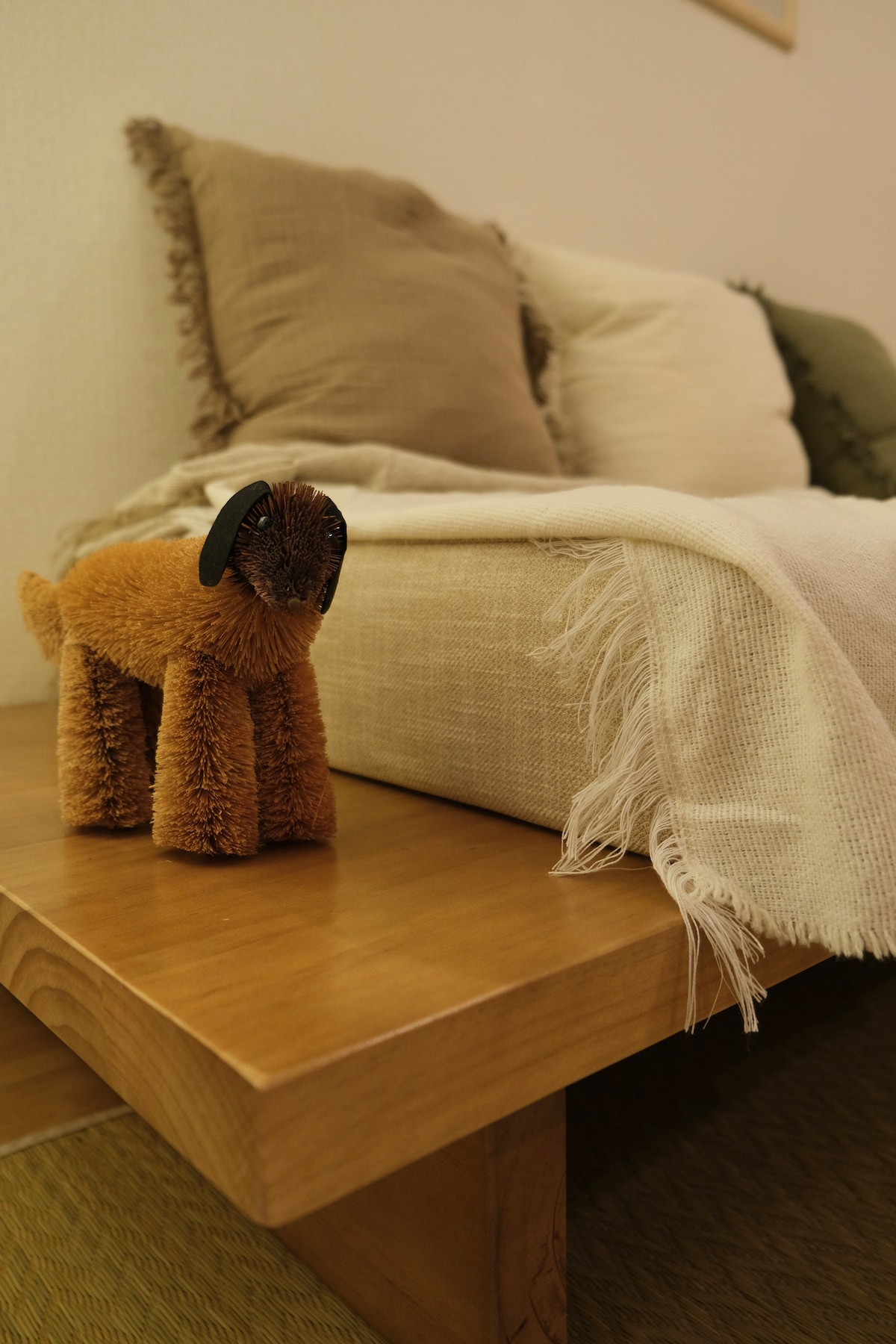 A close-up of a wooden table is featured, showcasing a charming plush dog toy and soft textiles. A cozy sofa with neutral-colored cushions is arranged near the table, with a light throw blanket casually draped across the seating area.