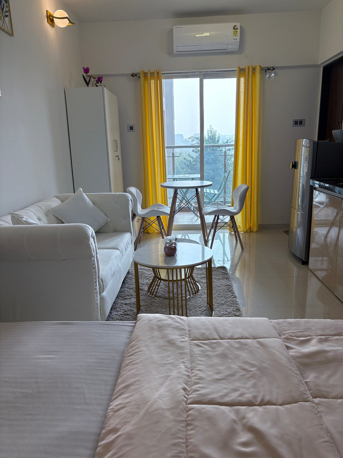 A light-filled living area features a comfortable white sofa, a dining table with two chairs, and large windows adorned with bright yellow curtains. A small round coffee table sits in front of the sofa, and a refrigerator is visible in the corner.
