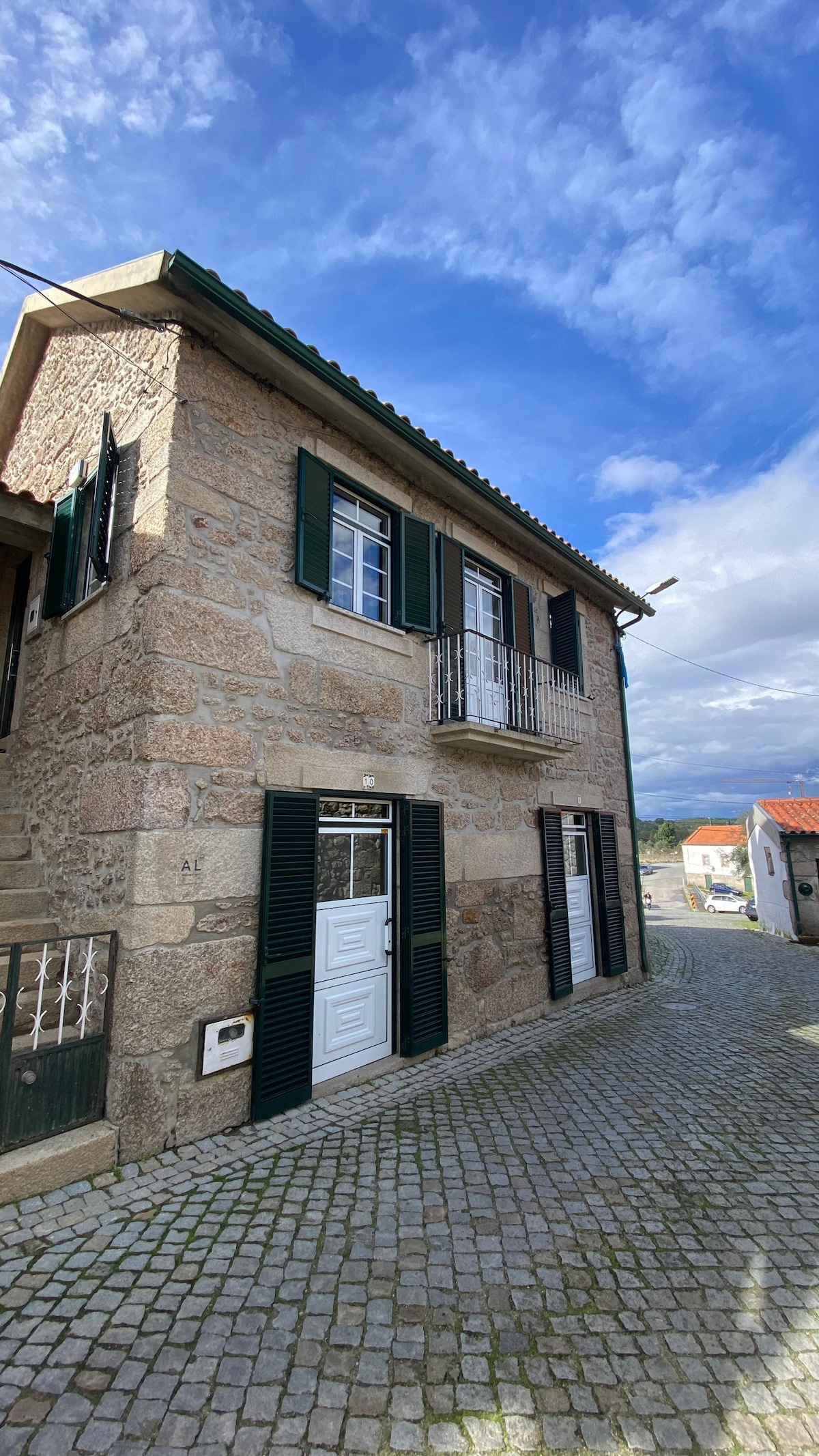 The exterior of the rustic house is characterized by its natural stone walls and dark green shutters. A small balcony is visible on the upper floor, adding charm to the facade. Cobblestone pathways lead up to the entrance, surrounded by a quiet village setting.