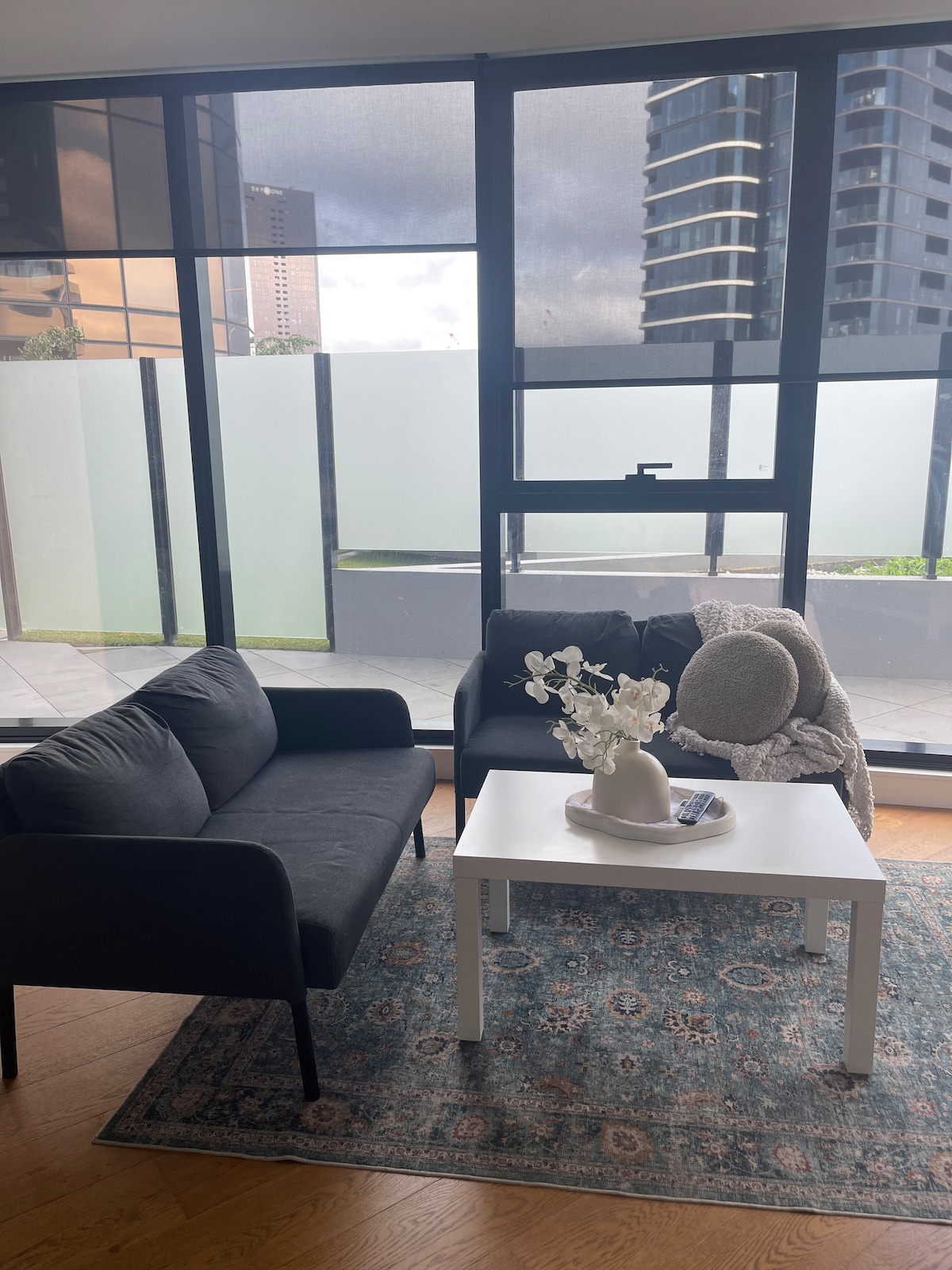 A modern living area features two dark sofas positioned beside a white coffee table. A vase with white flowers and a decorative pillow can be seen on the sofa. Large windows allow natural light to fill the space, while a patterned rug adds warmth to the wooden floor.