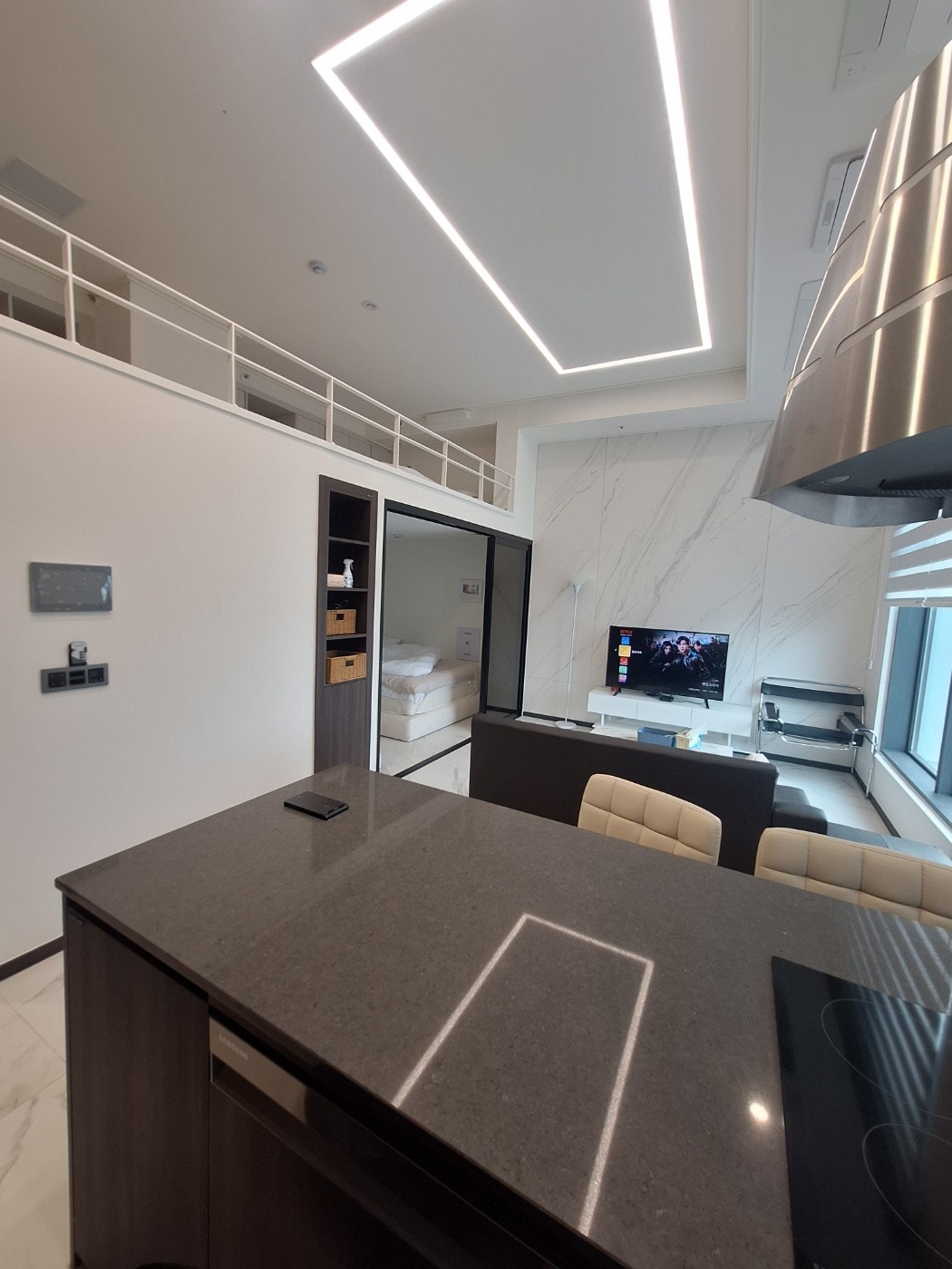An open interior space features a modern kitchen island with a polished countertop. A television is seen mounted on the wall opposite a seating area with light-colored chairs. The room is illuminated by recessed lighting, enhancing the sleek design and spacious feel.