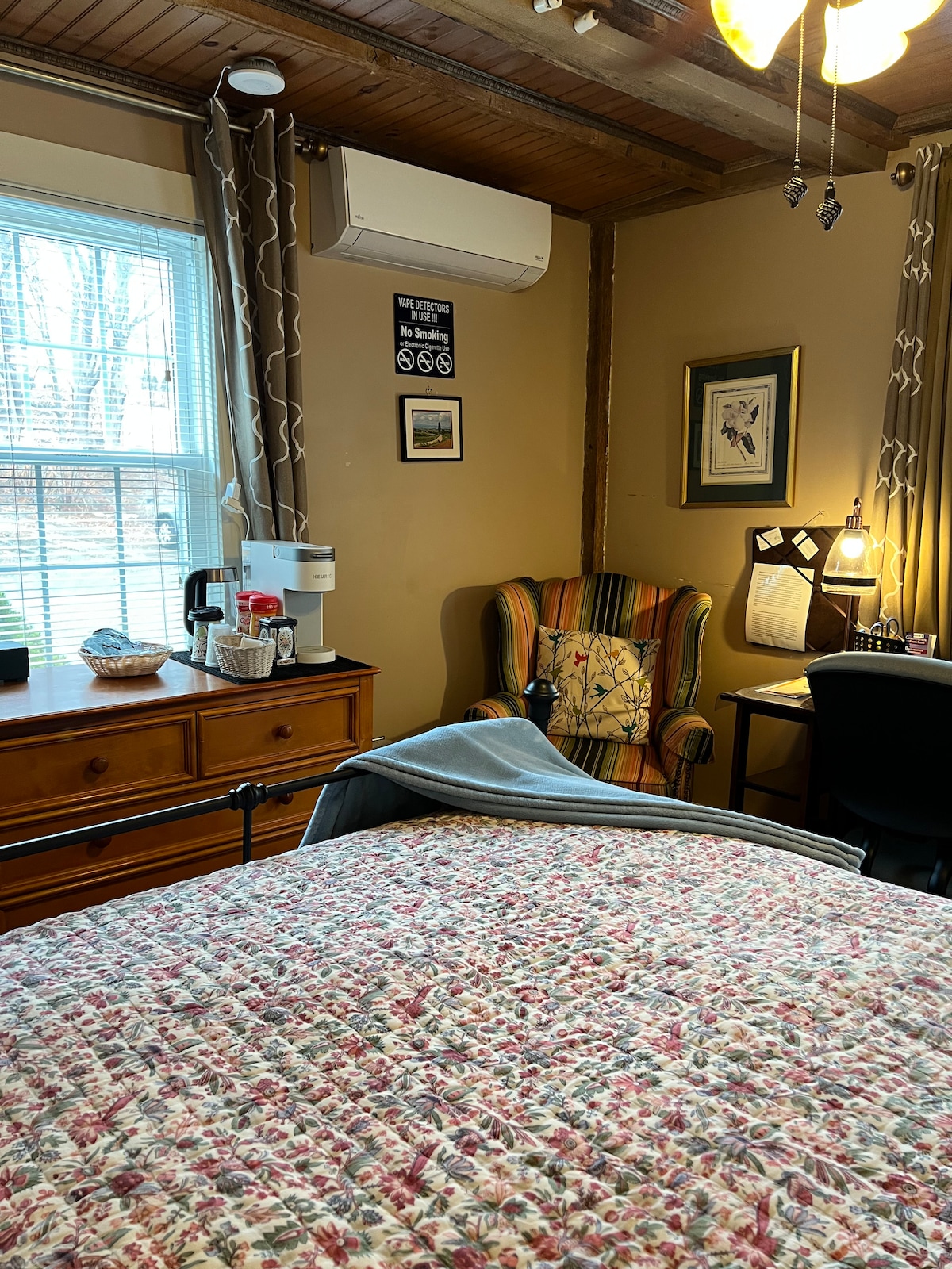 A comfortable room features a full size bed with a floral quilt. In one corner, a reading chair displays a colorful striped pattern. A desk with a lamp is positioned nearby, alongside a dresser equipped with a coffee maker and other amenities. Natural light filters through a window adorned with curtains.