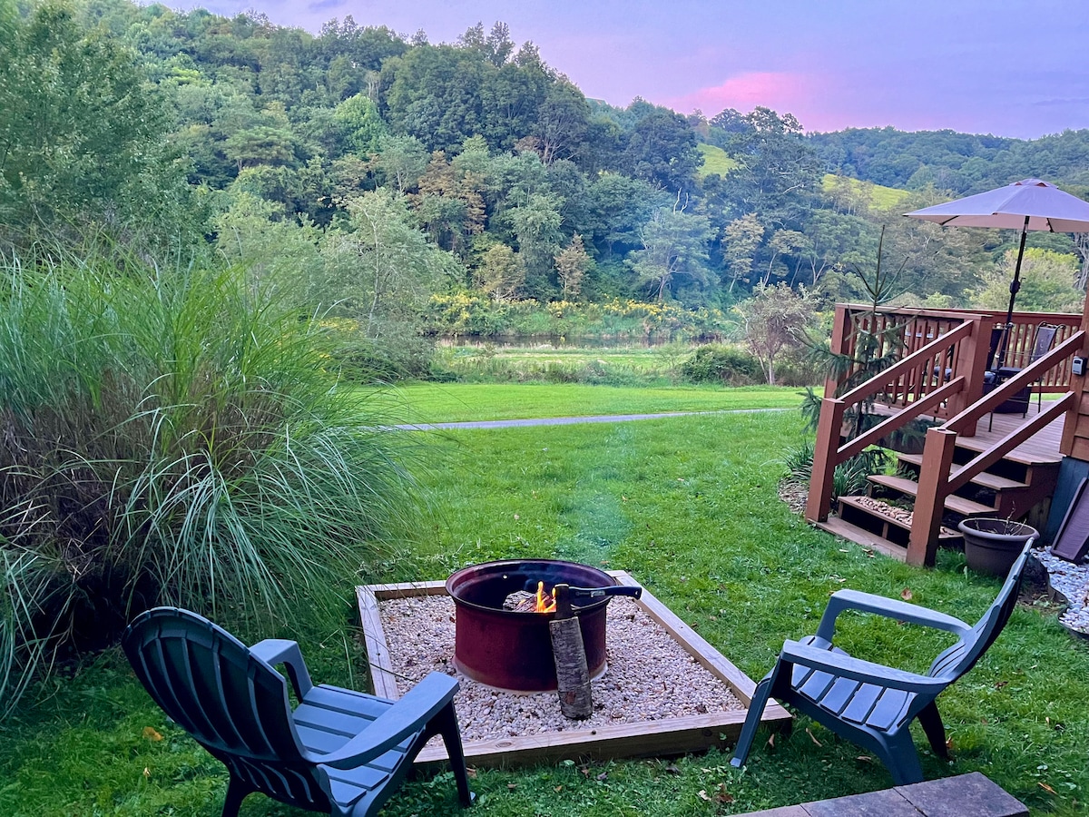 An outdoor fire pit, surrounded by gravel, is positioned on a grassy lawn, inviting relaxation. Two dark blue chairs face the flames. Lush greenery and distant hills are visible, bathed in soft, natural light. A wooden deck extends to the right, enhancing the outdoor space.