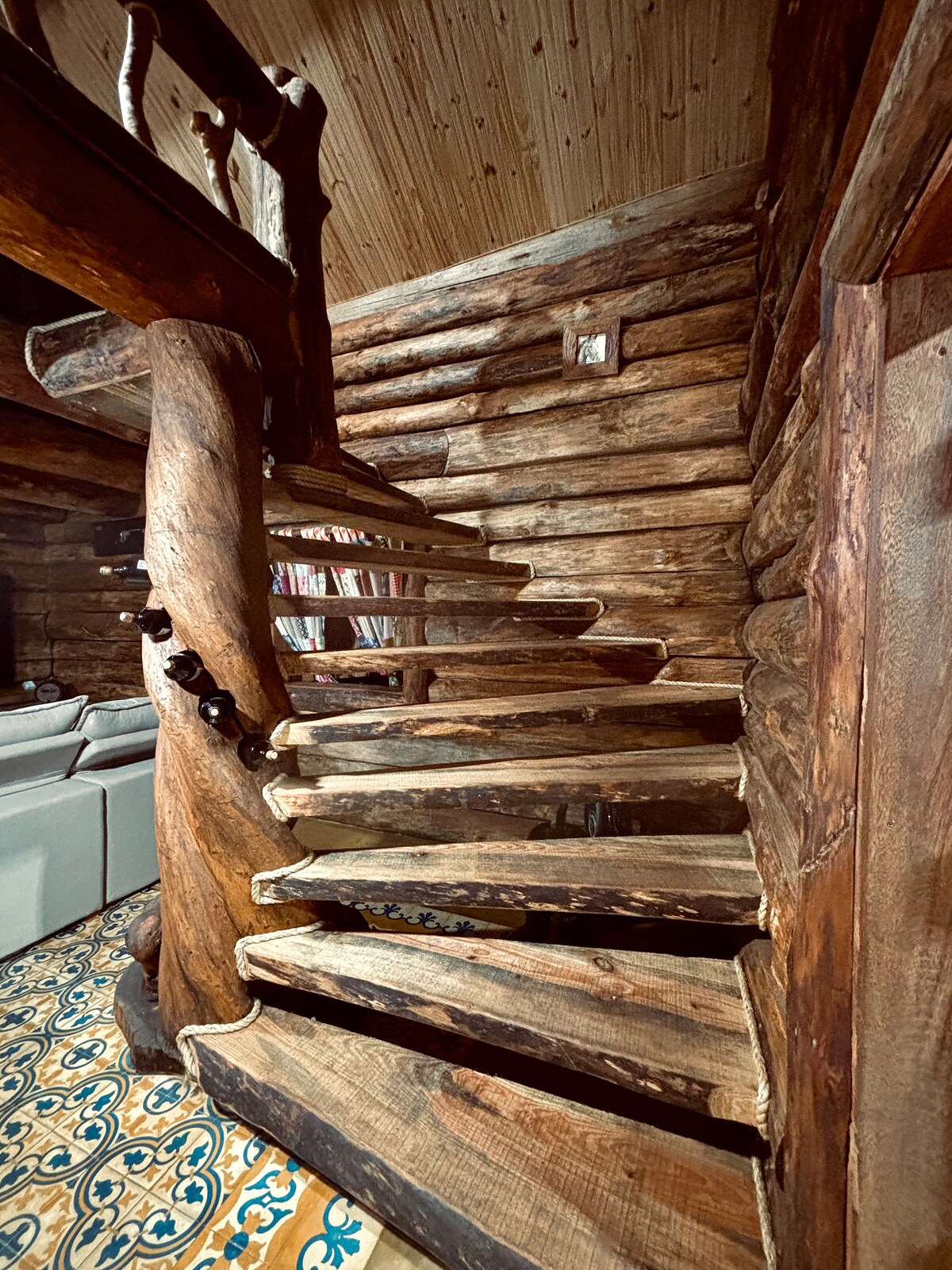 A spiral staircase crafted from rustic wood leads upwards, surrounded by log walls that emphasize the cabin's natural aesthetic. The sturdy railing blends seamlessly with the organic materials, while decorative patterned tiles are visible on the floor below.