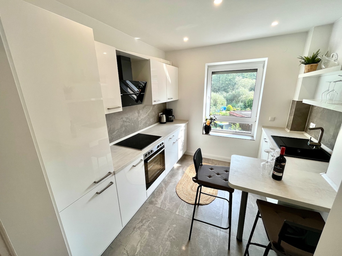 A modern kitchen features sleek white cabinetry and a gray countertop. A window offers natural light and a view of greenery. The space includes a built-in oven, cooktop, and refrigerator. A small dining table with chairs is arranged for cozy meals.