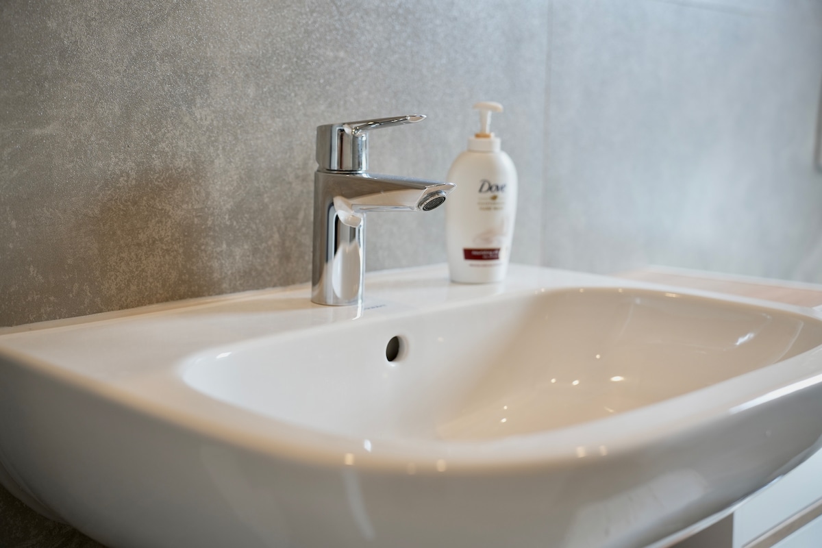 A modern sink is shown with a sleek chrome faucet and a smooth white basin. A bottle of Dove hand soap sits beside the sink on a light-colored countertop, contributing to a clean and functional bathroom space.