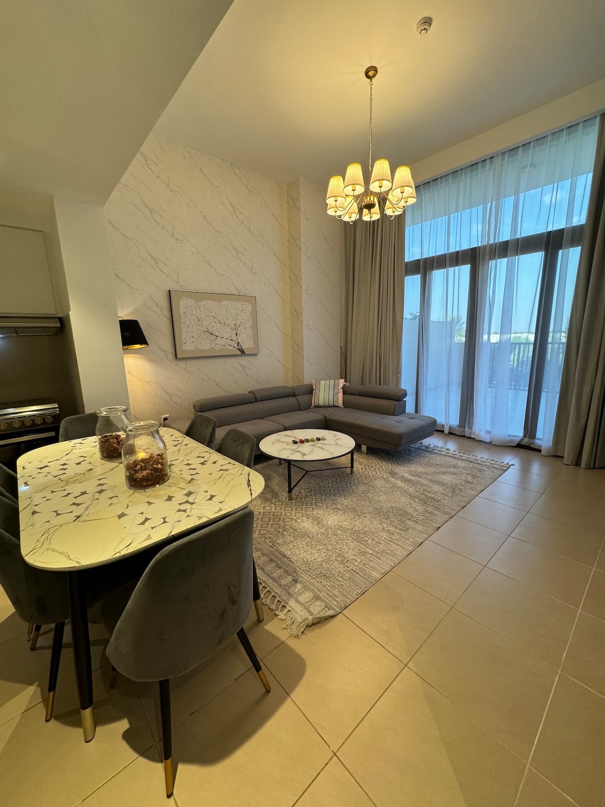 A well-lit living area features large windows that allow natural light to fill the space. A modern, neutral-colored sofa is positioned beside a round coffee table, while a dining table with plush chairs occupies the area. Elegant lighting fixtures hang from the ceiling.