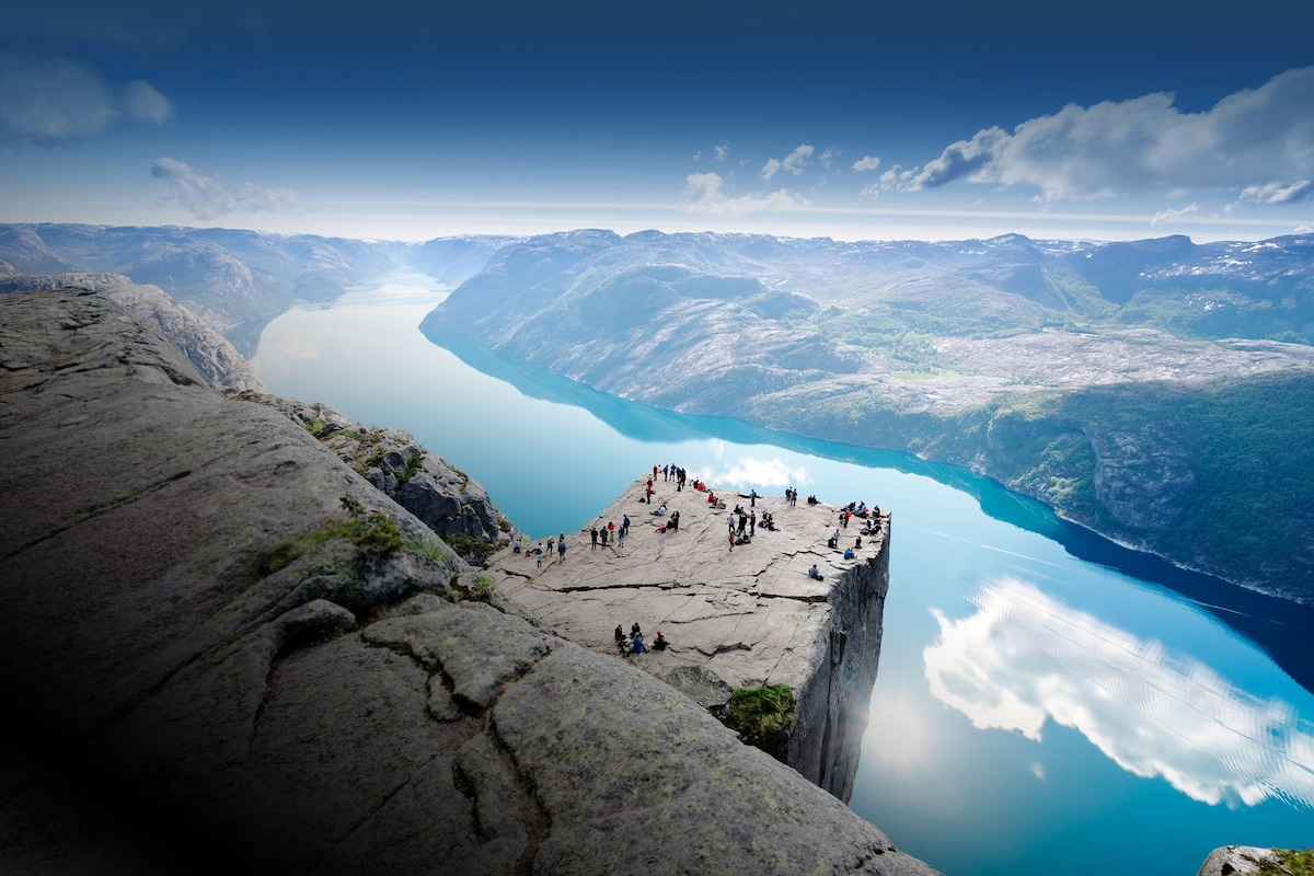 A vast rocky outcrop extends over a tranquil fjord, with a panoramic view of the water and surrounding mountains. Several individuals are positioned at the edge, illustrating the natural beauty of the landscape under a clear sky.