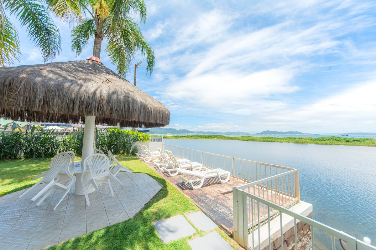 A shaded gazebo with a thatched roof is positioned next to a tranquil lagoon. Loungers are arranged around the outdoor space, with lush greenery framing the scene and distant hills visible under a clear blue sky.