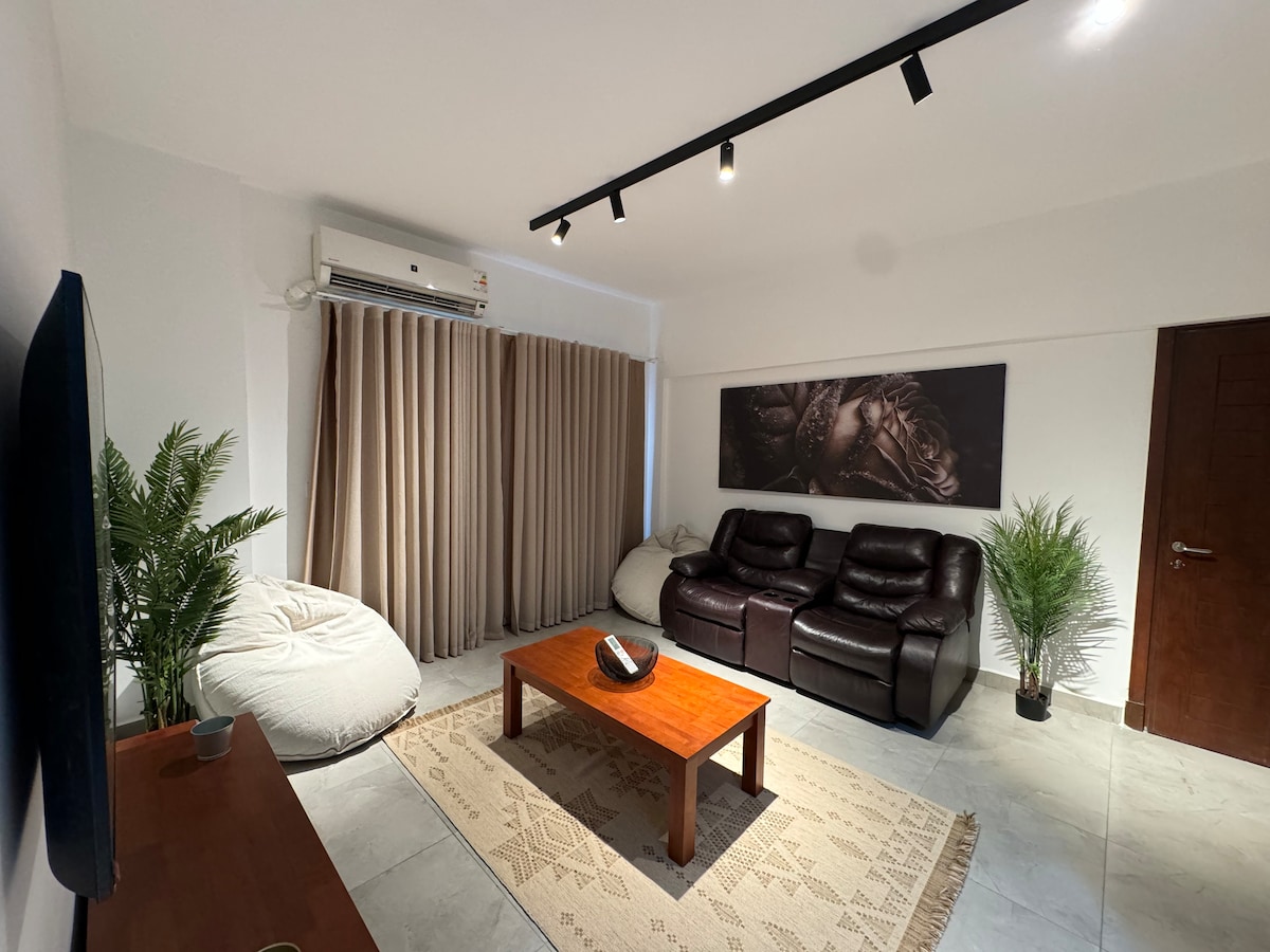The living area features a comfortable seating arrangement with dark leather recliners and large bean bags. A wooden coffee table sits at the center, surrounded by decorative plants. Soft natural light filters through floor-length curtains, enhancing the modern aesthetic.