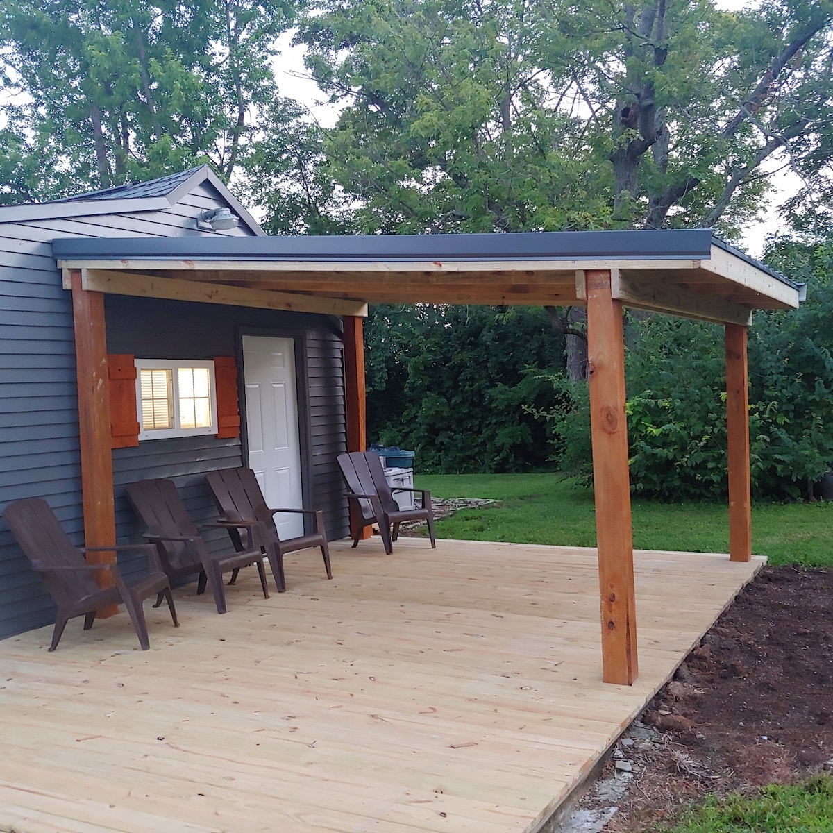 A partially covered deck extends from the exterior of the home, featuring several wooden adirondack chairs arranged for relaxation. A white door and two windows with orange shutters are visible, surrounded by lush greenery and trees in the background.