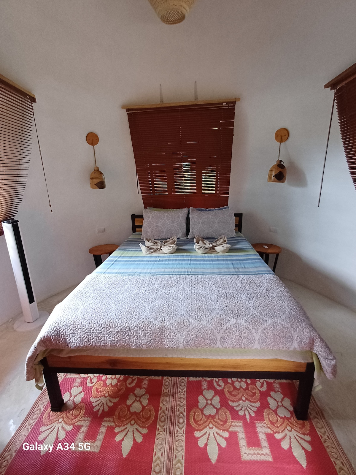 A bright bedroom features a spacious bed adorned with a textured blanket and coordinating decorative pillows. Wooden side tables are positioned on either side of the bed, and light filters through bamboo blinds above the windows, enhancing the serene setting.