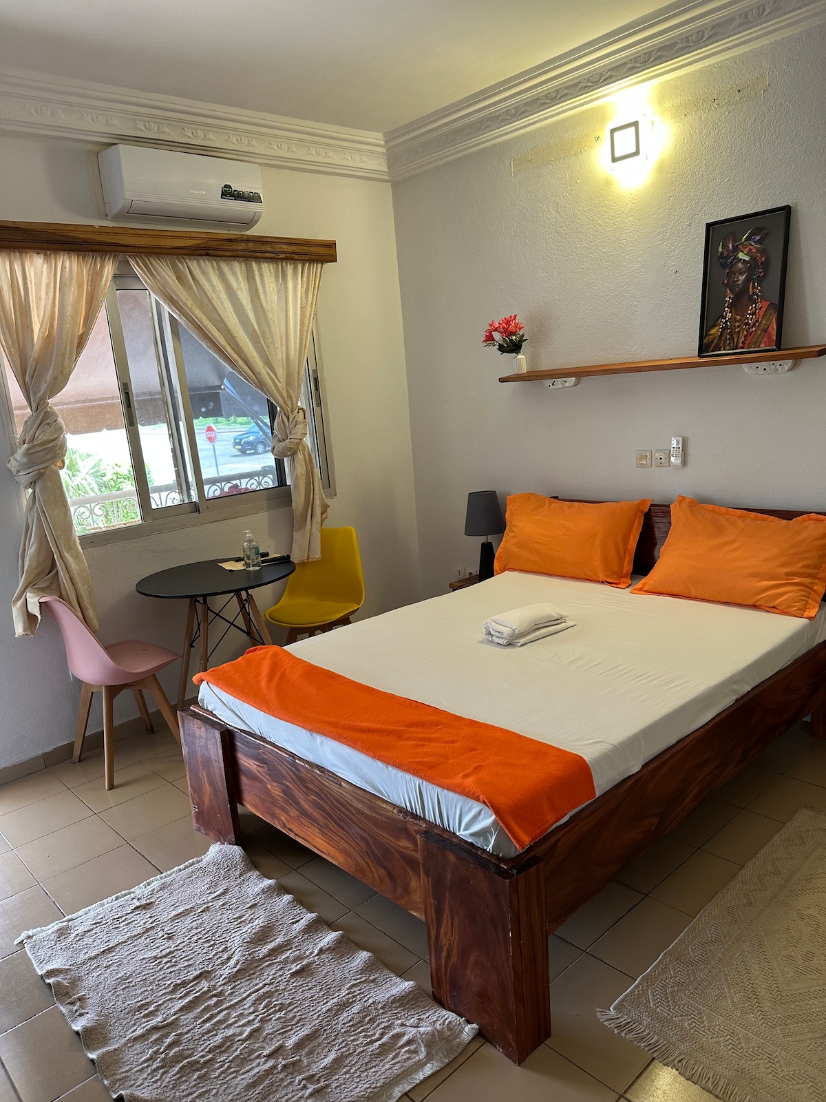 A well-lit bedroom features a spacious bed adorned with bright orange and white bedding. A small wooden table with two chairs is placed near the window, which is framed by soft curtains. Decorative elements include wall art and a small shelf with flowers.