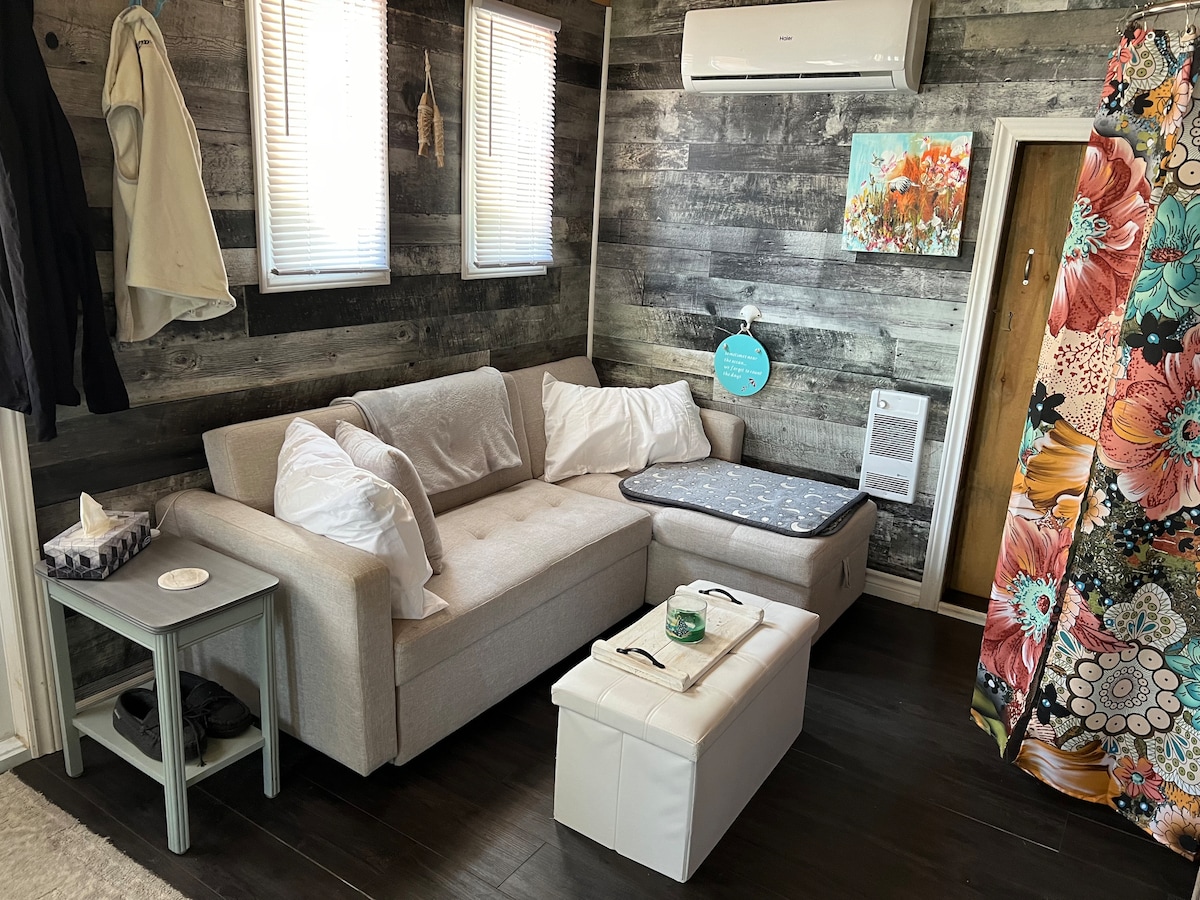 A light-colored sectional sofa with soft white cushions is positioned in a cozy corner. Above, two small windows allow natural light to brighten the rustic wood-paneled walls. A small side table and a white storage ottoman complement the seating area, enhancing the cabin's inviting design.