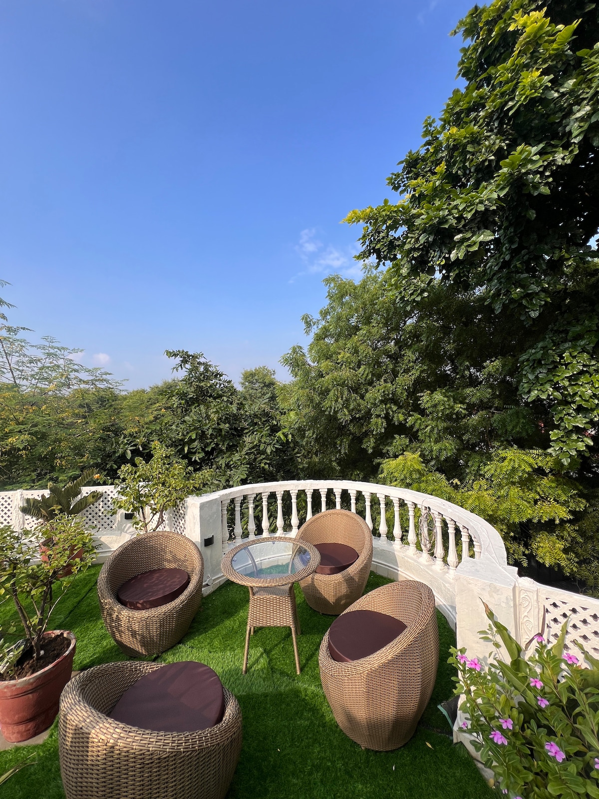 An outdoor terrace garden is visible, featuring two wicker chairs and a glass-top table. Lush greenery surrounds the area, providing a serene atmosphere. Flower pots add a touch of color, contrasting with the bright blue sky overhead.