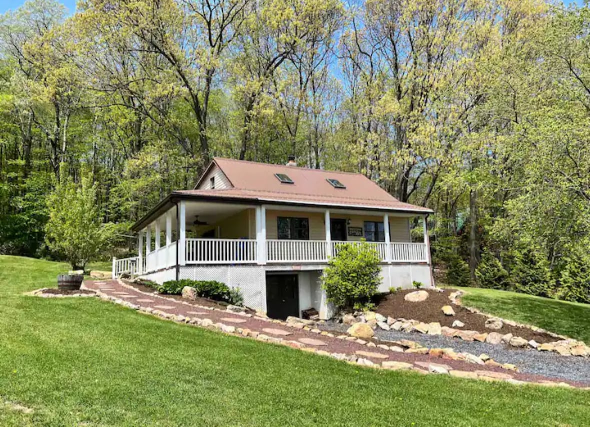 A charming two-story home is surrounded by vibrant greenery and colorful landscape. The covered front porch invites relaxation, while large windows allow natural light to fill the interior. A gently sloping yard leads to a well-maintained garden area, enhancing the peaceful outdoor setting.