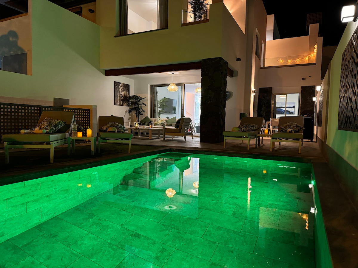 A heated swimming pool is illuminated in vibrant green, reflecting the surrounding outdoor area. Comfortable lounge chairs are positioned nearby, complemented by decorative elements. The villa's interior is visible through expansive windows, showcasing an inviting atmosphere with warm lighting.