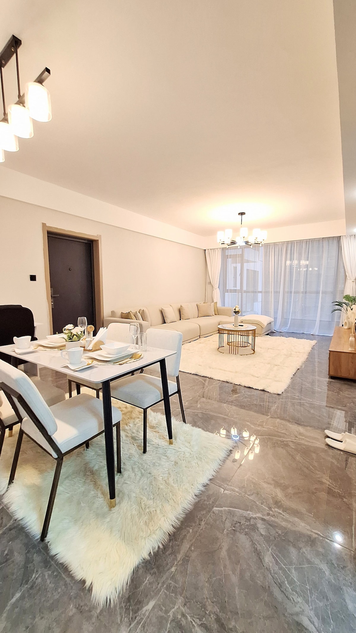 A modern living area features a plush beige sofa arranged around a coffee table, complemented by a light rug. A dining table with white chairs is set nearby, creating an inviting space for meals. Large windows allow natural light to brighten the room.