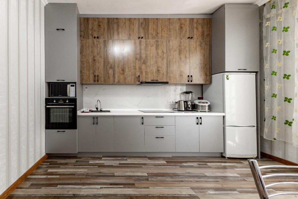 A modern kitchen area is equipped with sleek cabinetry in a two-tone finish, featuring wooden accents. Essential appliances are visible, including an oven and refrigerator. Light-colored walls complement the wood flooring, while a window adorned with sheer curtains allows natural light to enter.