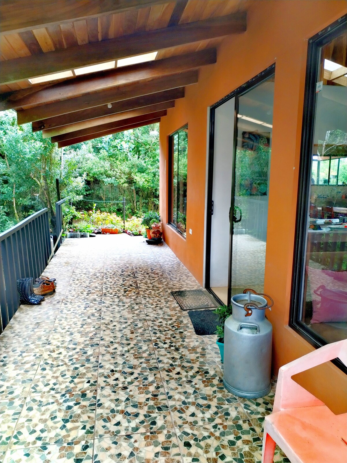 An inviting porch features a tiled floor and wooden beams overhead. Large windows offer views of the lush greenery outside, with flower beds visible in the distance. A small gas cylinder and a plastic chair are positioned along the side, enhancing the practical outdoor space.