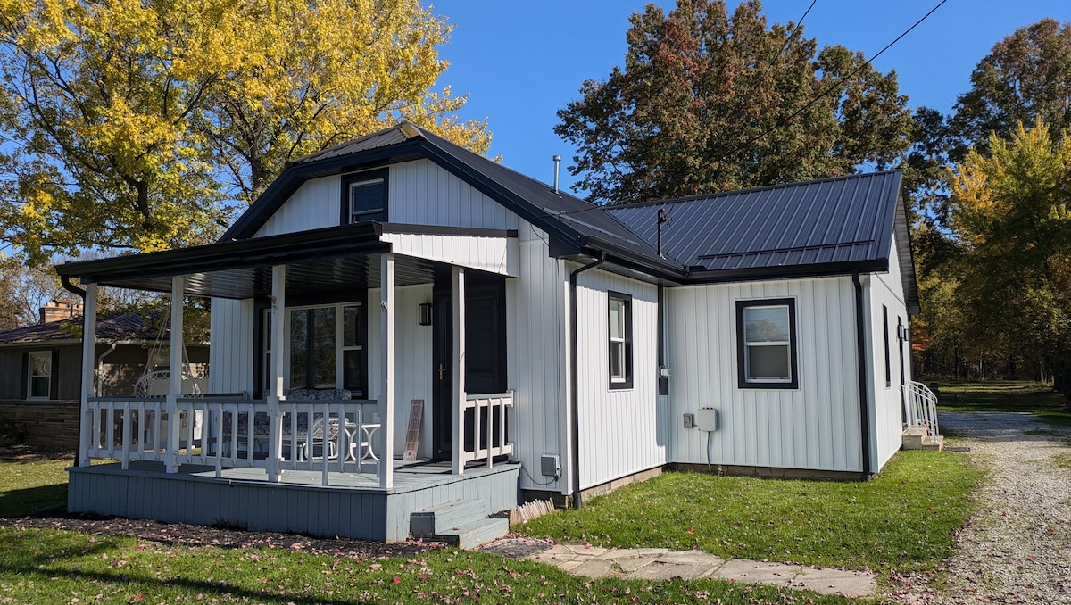 The exterior of the house features a welcoming porch with white railings. The dwelling is painted in white with black accents, showcasing a metal roof. A lawn surrounds the property, bordered by trees displaying autumn foliage.