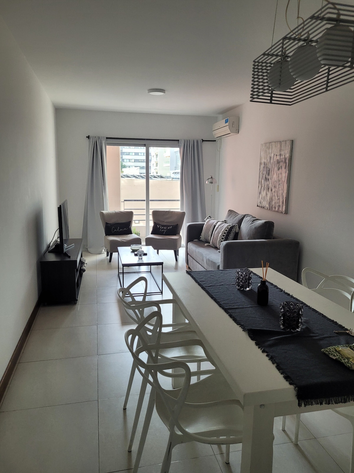 A bright living area features a comfortable gray sofa and two armchairs beside a coffee table. A dining table with six chairs is visible, and a wall-mounted TV is placed across from the seating. Large sliding glass doors lead to a balcony, allowing natural light.