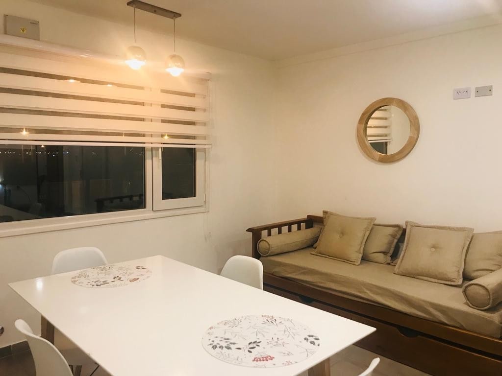 A bright dining area features a white table surrounded by modern chairs. A cozy bench with plush cushions is positioned against the wall beneath a circular mirror, while soft light from pendant fixtures casts a warm glow across the space.