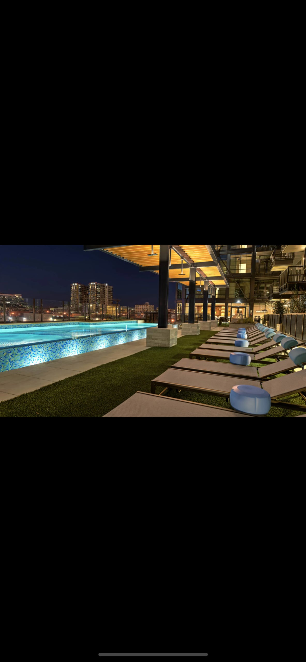 The rooftop lounge area features a shimmering pool side by side with lounge chairs. Soft lighting enhances the modern design, while city views illuminate the skyline during the night, creating a serene atmosphere for relaxation and socializing.