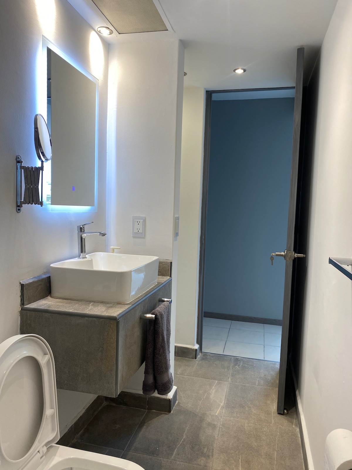A modern bathroom features a sleek sink with a minimalist design and a large mirror above. Soft lighting creates a bright atmosphere. A towel hangs from a bar beside the toilet, and the floor is tiled with dark stone, complementing the overall contemporary aesthetic.