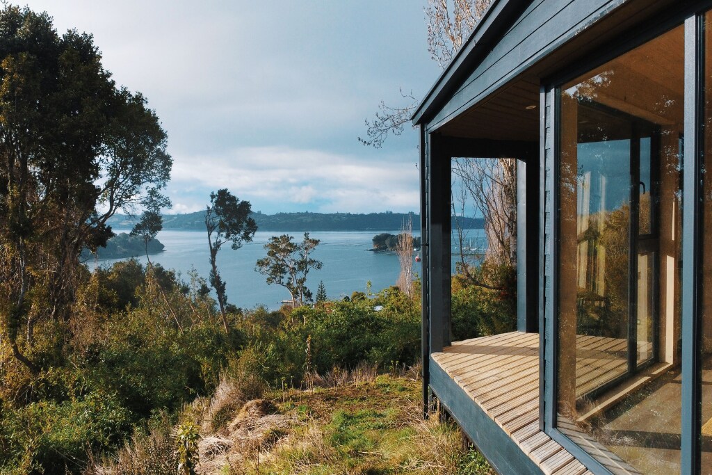 A panoramic view of the serene coastline is offered from the deck of the house, framed by large glass windows. Lush greenery surrounds the property, enhancing the connection to nature and the peaceful ambiance.