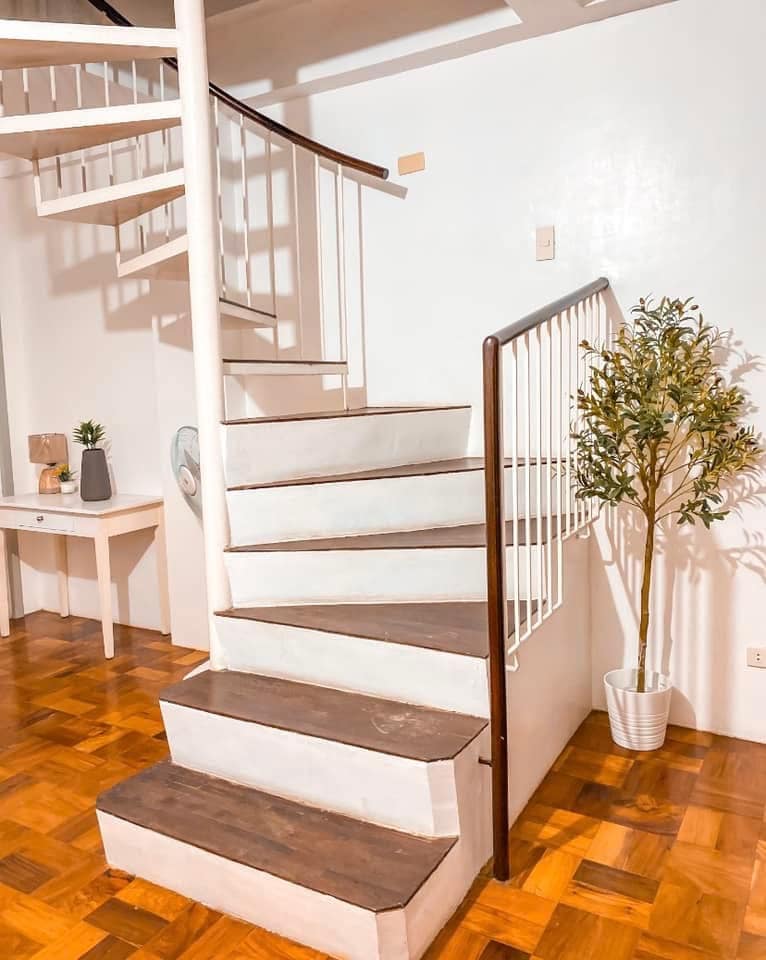 A spiral staircase leading to the upper level is showcased, featuring white steps with brown accents. A small decorative plant in a white pot is positioned beside the staircase, contributing to the minimalist decor of the space. The warm wooden floor adds a cozy touch.