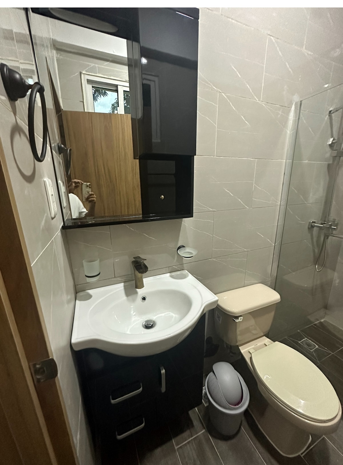 The bathroom features modern fixtures with a glass shower enclosure, a white vanity with a sink, and a toilet. Storage cabinets are mounted above the sink. Natural light enters from a window, illuminating the tiled walls and floor.