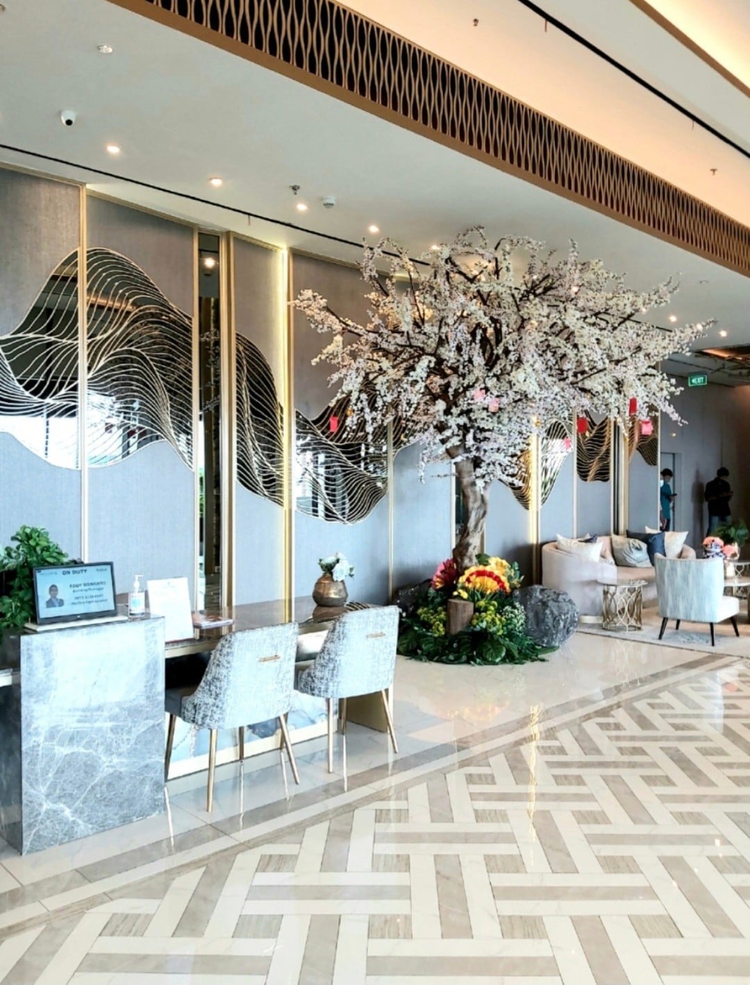 A spacious lobby features elegant marble flooring and modern decorative elements. A large cherry blossom tree adds a natural touch, accompanied by stylish seating options. The walls are adorned with abstract designs, creating a welcoming and contemporary atmosphere for guests.