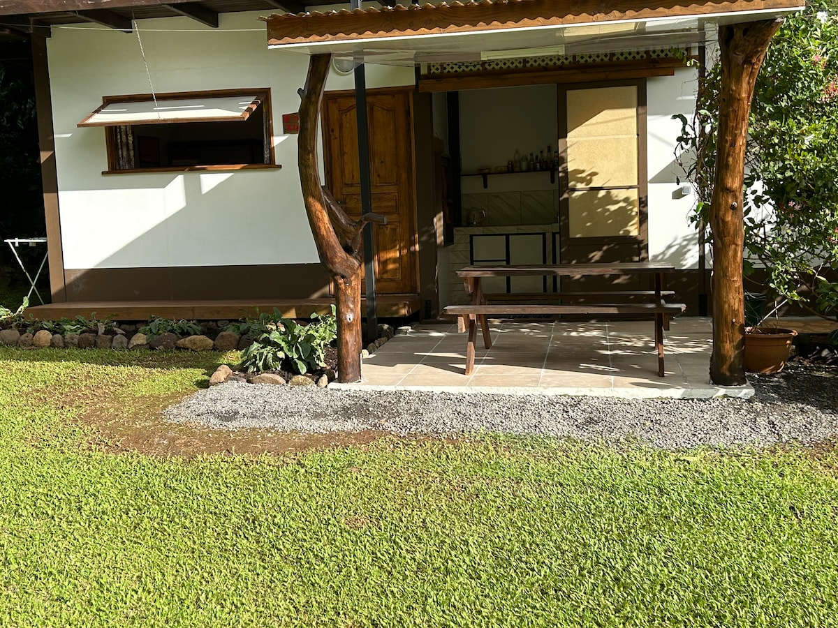 A covered outdoor seating area is presented with a wooden table and benches, surrounded by a grassy lawn. The entrance features a rustic wooden door and a small window, framed by lush greenery and stone accents.