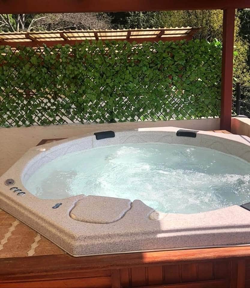 A spacious jacuzzi is situated under a wooden gazebo, surrounded by lush greenery. The bubbling water creates a relaxing ambiance, and the textured surface of the jacuzzi is visible, offering an inviting space for relaxation.