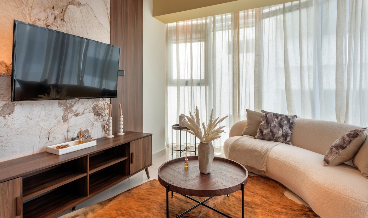 A modern living area features a large, curved sofa and a wooden coffee table. A flat-screen television is mounted on the wall, while natural light filters through sheer curtains, creating a soft glow in the space. Decorative elements, including a vase with dried plants, add a touch of elegance.