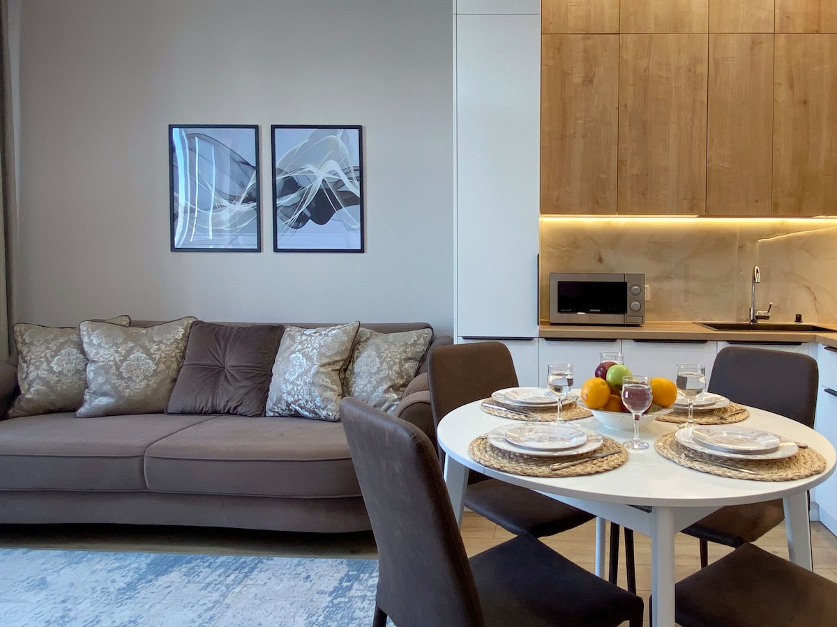A comfortable living area features a plush brown sofa adorned with decorative pillows, complemented by a dining table set for four. Behind, modern cabinetry houses kitchen appliances. Soft lighting enhances the space, while art pieces are displayed on the walls.