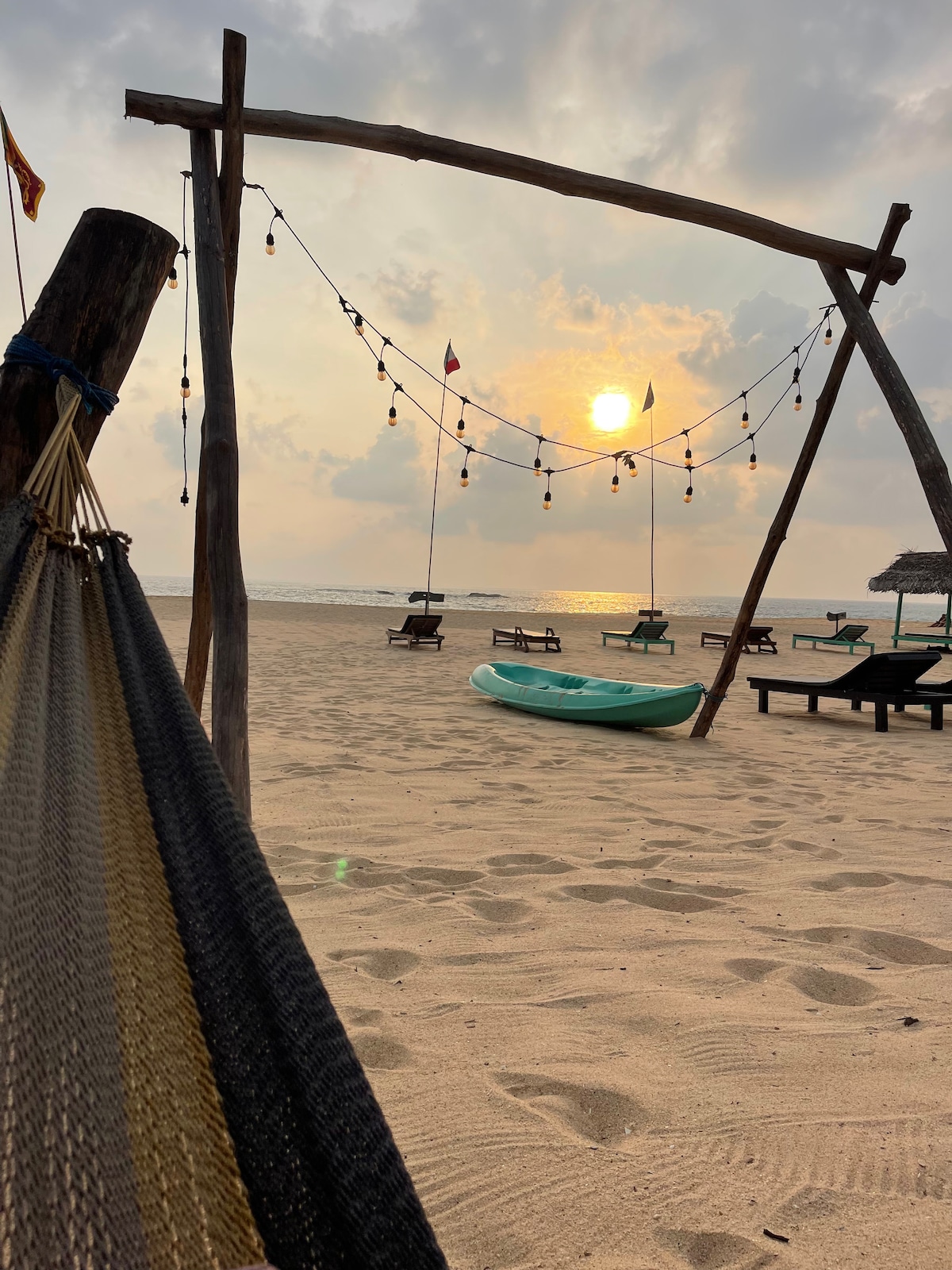 A gently swaying hammock is seen in the foreground, with the sun setting on the horizon. Beach loungers are arranged on the sand, and decorative lights hang from a wooden frame. The calm sea reflects the warm colors of the evening sky.