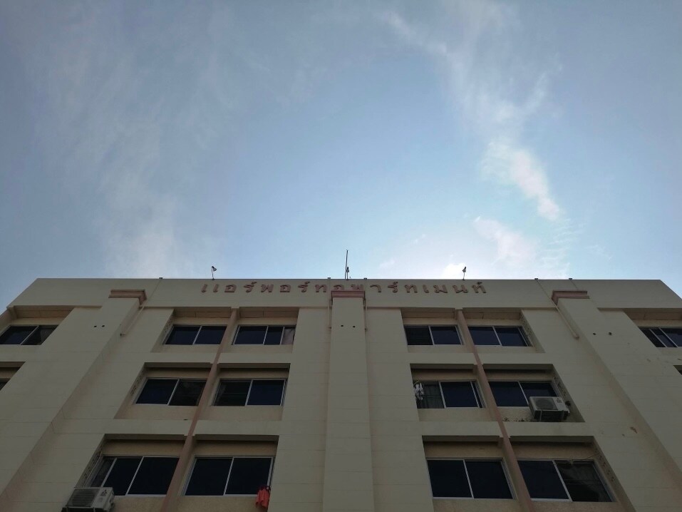 The exterior of a multi-story building is captured from a low angle, showcasing large windows and air conditioning units. The sky above appears clear with a few wispy clouds, and the building's name is displayed prominently at the top.