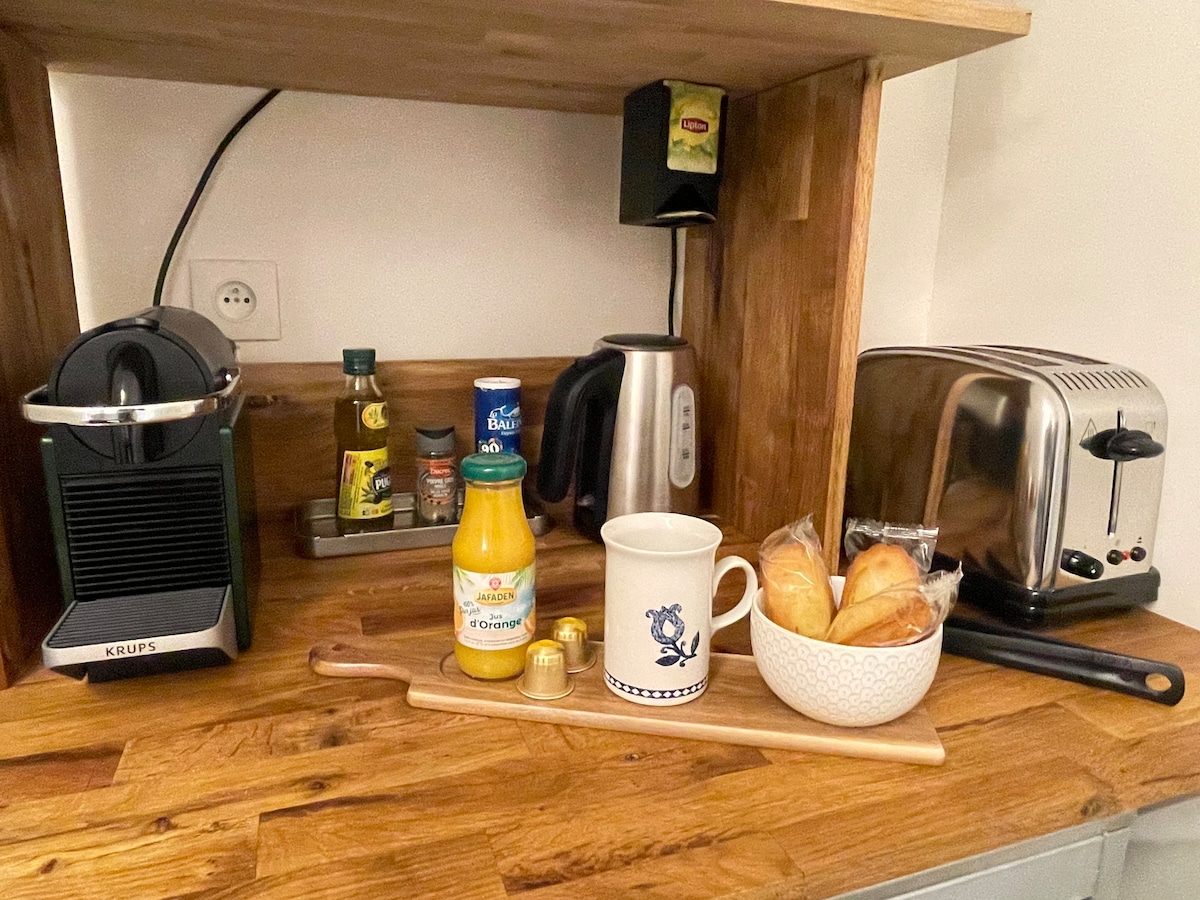 The kitchenette is designed with wooden shelving, featuring a coffee machine, an electric kettle, and a toaster. A mug and a container of orange juice sit alongside a small bowl of bread rolls. Various condiments are arranged on the shelf, adding functionality.