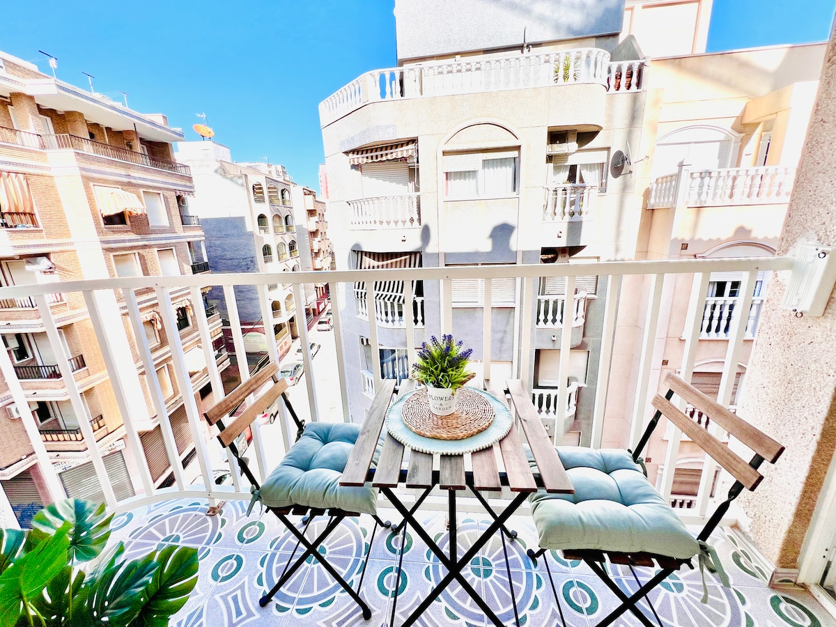 A small balcony features two folding chairs and a table, which is adorned with a decorative plant. The view reveals surrounding buildings, showcasing a mix of architectural styles. Bright sunlight illuminates the space, enhancing the vibrant floor tiles below.