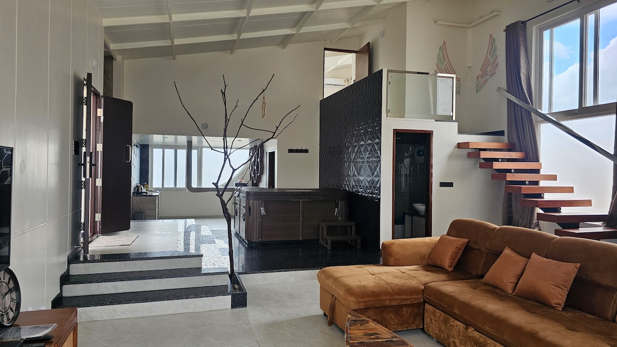 A spacious living area is highlighted by a large, brown sectional sofa with several decorative pillows. Light streams through expansive windows, illuminating the stylish décor. A sleek staircase leads to an upper level, while a bar counter is visible in the background, enhancing the open layout.