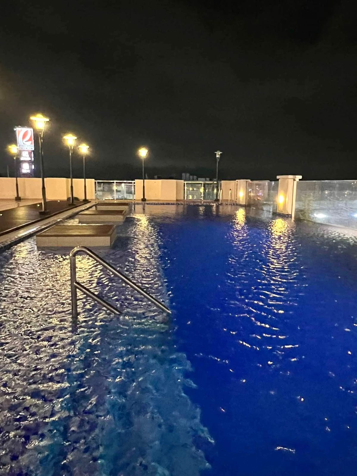 An illuminated swimming pool is shown at night, with clear blue water reflecting the lights from surrounding lampposts. The area features steps leading into the pool and a glass barrier for safety, creating a serene atmosphere.
